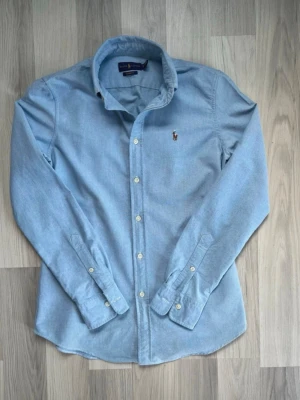  Ralph Lauren skjorta - Klassisk ljusblå skjorta från Ralph Lauren med knappstängning framtill och broderad logga på bröstet. Skjortan har lång ärm, krage och är tillverkad i mjuk bomull. Perfekt för en stilren och avslappnad look.