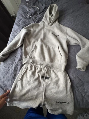 Grå Essentials hoodie & shorts set - Säljer ett grått mjukisset från Essentials i storlek Xs men jag själv har M och passar perfekt alltså är det over sized . Setet består av en hoodie med känguruficka och dragsko samt matchande shorts med resårmidja och snörning. Båda plaggen har Essentials-logga tryckt framtill. Perfekt för chill dagar hemma.