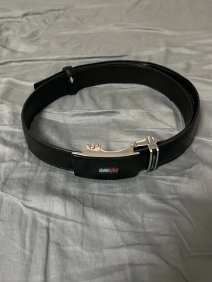 Svart Tommy hilfiger bälte med metallspänne - Snyggt svart Tommy hilfiger  bälte i slätt läder med ett stilrent, blankt metallspänne. Bältet har en modern design och passar perfekt till jeans eller kostymbyxor. Spännet har en unik form som ger ett exklusivt intryck. Priset kan diskuteras vid snabb affär.