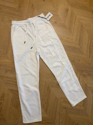 Vita mjukisbyxor från Define - Snygga vita mjukisbyxor från Define med resår i midjan och dragsko. Byxorna har sidofickor och en stor vit logga längs benet för en trendig look. Perfekta för chill dagar eller när du vill ha en clean stil., skriv för frågor om passstorleken!