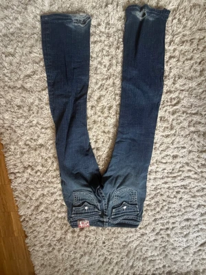 Blå bootcut jeans med slitningar - Säljer ett par blå bootcut jeans med slitningar och snygga detaljer på bakfickorna. Jeansen har ljusare partier framtill och grova sömmar. Klassisk femficksmodell i denim med låg midja och dragkedja. Perfekta för en avslappnad och trendig look.