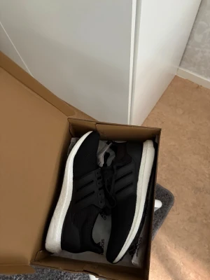 Svarta Adidas Ultraboost sneakers - Svarta Adidas Ultraboost sneakers med ventilerande mesh-ovandel, klassiska tre ränder på sidan och vit Boost-mellansula för extra komfort. Yttersulan är svart med greppvänligt mönster och en grön detalj i sulan. Perfekta för både streetstyle och träning.