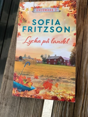 Lycka på landet av Sofia Fritzson - En romantisk och charmig pocketbok av Sofia Fritzson, del tre i serien om byn Ängsmark. Omslaget har höstfärger med gula och orange löv, en lantlig gård och paraply. Perfekt för dig som gillar feelgood och idylliska landsbygdsskildringar.