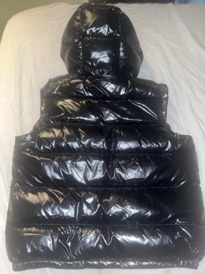 Moncler väst - Snygg och stilren Moncler-dunjacka i svart med klassisk blank finish. Varm och perfekt för höst/vinter. Jackan har huva, dragkedja framtill, två praktiska sidofickor och den klassiska Moncler-loggan på bröstet.