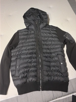 Svart pufferjacka med huva Moncler - Snygg svart pufferjacka från Moncler med huva och stickade ärmar. Jackan har två fickor med tryckknappar och en Moncler-logga på vänster ärm. Perfekt för kalla vinterdagar och ger en sportig look.