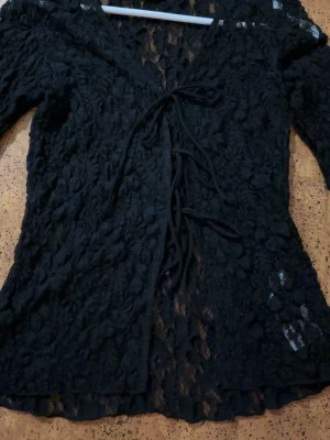 Svart spetskofta från Gina Tricot - Säljer en svart, långärmad spetskofta från Gina Tricot. Koftan har knytning framtill och är tillverkad i ett genombrutet, blommigt spetsmaterial som ger en trendig och luftig look. Perfekt att styla över ett linne eller topp.