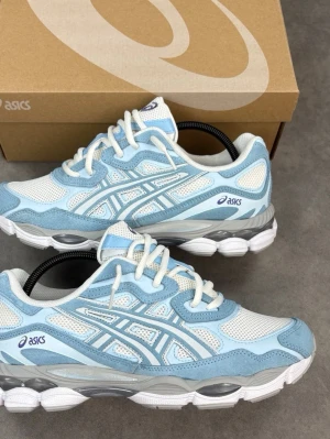  Nya Asics  nyc gel sneakers - Snygga sneakers från Asics i en fräsch ljusblå och vit färgkombination. Ovandelen är i mesh och mockaliknande material med klassiska Asics-stripes på sidorna. Skorna har vita snören och en chunky vit och grå sula som ger en sportig vibe.