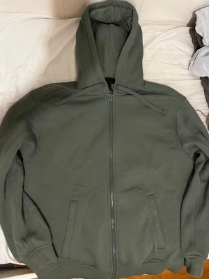 Olivgrön hoodie med dragkedja - Säljer en olivgrön hoodie med dragkedja framtill och två fickor. Tröjan har huva med dragsko och ribbade muddar vid ärmar och nederkant. Perfekt för en chill och avslappnad stil. Materialet känns mjukt och bekvämt, troligen bomullsblandning.