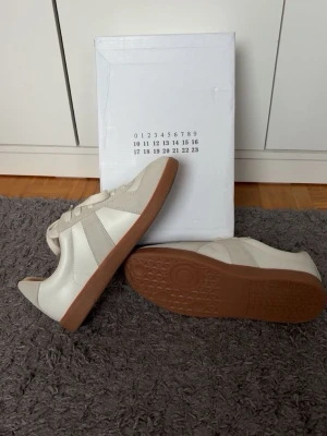 Vita maison margiellas - Stilrena sneakers i vitt och beige med klassisk låg siluett. Ovandelen är i en mix av slätt läder och mocka, och sulan är i gummi med brunt greppmönster. Skorna har vita snören och en tidlös design som passar till det mesta. Storlek 42. Helt nya 