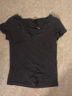 Brun topp med spetsdetalj - Mörkbrun topp med v-ringning och dekorativ spets längs halsringningen. Kortärmad och i mjukt material som känns skönt mot huden. Perfekt för dig som gillar enkla men snygga detaljer.