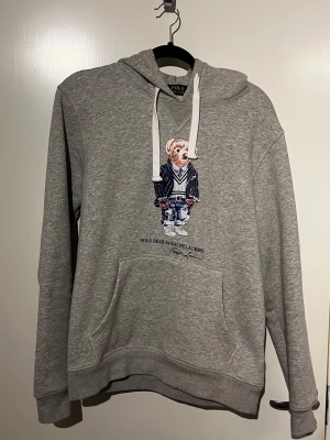 Grå hoodie Polo Bear Ralph Lauren - Grå hoodie med huva och vit dragsko från Ralph Lauren. På bröstet finns ett stort tryck av den ikoniska Polo Bear i preppy outfit. Hoodien har känguruficka och är gjord i mjukt material, perfekt för en chill och stilren look.