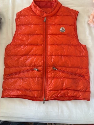 Orange dunväst från Moncler - Snygg röd dunväst från Moncler med hög krage och två dragkedjeförsedda fickor framtill. Västen har quiltad design och Moncler-logga på bröstet. Perfekt för lager-på-lager och ger en sportig vibe till din outfit. Size 2 för liten för mig tyvärr. Finns låda och kvitto från mister porter 