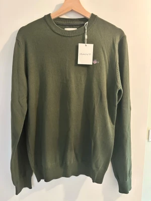 Mörkgrön GANT crewneck - Snygg mörkgrön crewneck från GANT med rund halsringning och diskret broderad logga på bröstet. Tröjan har långa ärmar och ribbade muddar vid ärmslut och nederkant. Perfekt till lager på lager och enkel att matcha.