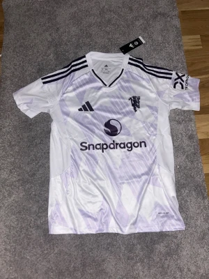 Vit Manchester United tröja från Adidas  - Snygg vit fotbollströja från Adidas med lila detaljer och tre klassiska ränder på axlarna. Tröjan har tryck med Snapdragon-logga och klubbmärke på bröstet. Tillverkad i lätt och ventilerande polyester, perfekt för träning eller match. S i storlek men sitter som M