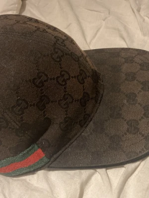 Brun keps med monogram och ränder - Snygg brun keps med mörkbrunt monogrammönster och klassiska gröna och röda ränder på sidan. Justerbar baktill för perfekt passform. Perfekt accessoar för dig som vill sticka ut med en trendig och tidlös look.