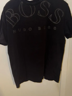 Svart t-shirt från Hugo Boss - Snygg svart t-shirt från Hugo Boss med stort BOSS-tryck och logga framtill. Klassisk passform och rund halsringning. Perfekt till jeans eller shorts för en clean och stilren look.