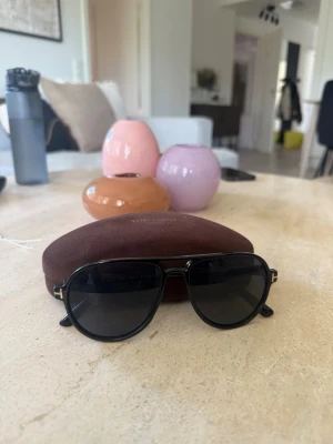 Svarta pilotbågar från Tom Ford - Snygga svarta solglasögon från Tom Ford med klassisk pilotform. Glasögonen har mörka linser och tunna bågar i metall. Kommer med originalfodral och ask, perfekt för dig som vill ha en stilren accessoar till sommaren.
