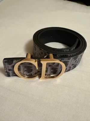Svart och grått bälte med guldspänne - Snyggt bälte i svart och grått med ett stort guldigt spänne med bokstäver. Bältet har ett mönster längs hela bandet och passar perfekt för att lyfta vilken outfit som helst.