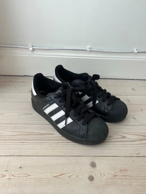 Adidas Superstar svartvita sneakers - Klassiska Adidas Superstar sneakers i svart och vitt med de ikoniska tre ränderna på sidan och texten 'SUPERSTAR'. Skorna har snörning, tåhätta i gummi och ovandel i läder. Endast använda i en månad. 