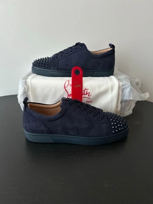 Mörkblå sneakers med nitar Louboutin - Snygga mörkblå sneakers från Christian Louboutin i mocka med svarta nitar på tån och klassisk röd sula. Skorna har lågt skaft, matchande mörkblå snörning och en exklusiv känsla. Perfekta för dig som vill sticka ut med stil. Allt OG medföljer förutom OG box