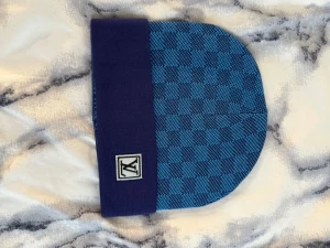 Blå mössa - Snygg mössa i blått med rutigt mönster och mörkblå uppvikt kant. På kanten sitter en patch med ett stilrent märke. Perfekt accessoar för att lyfta din outfit och hålla dig varm under kyliga dagar.