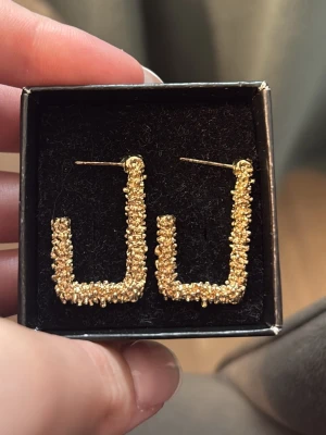 Guldiga fyrkantiga örhängen - Säljer ett par unika örhängen i guldton med fyrkantig form. Ytan är täckt av små, pärlliknande detaljer som ger en cool och trendig look. Perfekta för dig som vill sticka ut med accessoarer som syns. Oanvända 