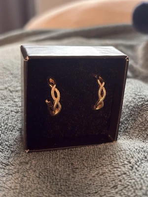 Guldiga tvinnade örhängen - Säljer ett par snygga örhängen i guldfärgad metall med tvinnad design. De har en elegant och modern look som passar perfekt till din stil. Kommer i en svart ask. Oanvända 