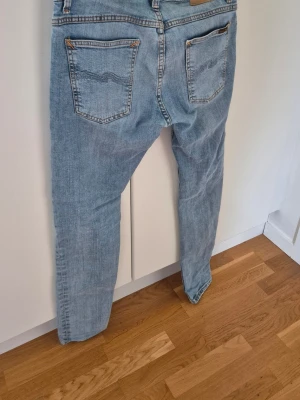 Nudie Jeans - Säljer ett par ljusblå jeans från Nudie med klassisk femficksmodell och raka ben. Jeansen har subtila slitningar och kontrastsömmar, samt Nudies ikoniska broderi på bakfickorna. Perfekta till en avslappnad stil och tillverkade i bomull.