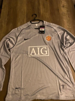 Manchester United grå långärmad tröja - Säljer en grå långärmad Manchester United-tröja från Nike med AIG-logga på bröstet. Tröjan har klubbmärket broderat, svart Nike-logga och är tillverkad i ett lätt, ventilerande material med NikeFIT-teknologi. Perfekt för fotboll eller träning.