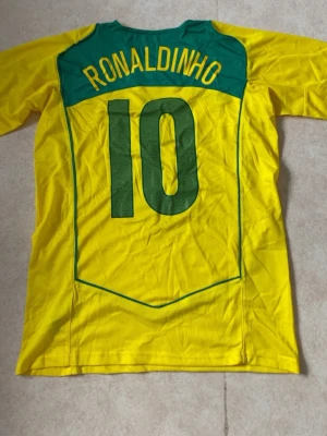 Brasilien Ronaldinho matchtröja gul - Gul Brasilien fotbollströja med gröna detaljer och nummer 10 på både fram- och baksida. Ronaldinho står tryckt på ryggen. Tröjan har korta ärmar, V-ringning och Nike-logga på bröstet. Tillverkad i lätt och ventilerande material, perfekt för fotboll eller träning.