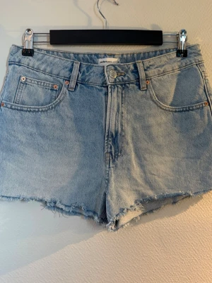 Ljusblå denimshorts med rå kant - Säljer ett par ljusblå denimshorts med låg midja och rå, fransig kant. Klassisk femficksmodell med knapp och dragkedja framtill. Perfekta till sommaren och ger en avslappnad vibe.
