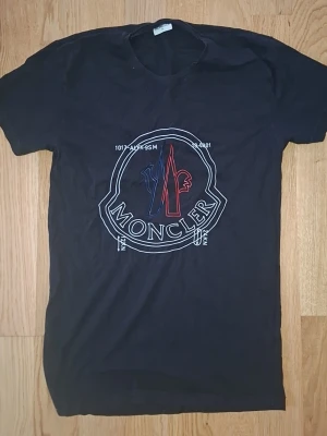 Svart Moncler t-shirt med tryck - Snygg svart t-shirt från Moncler med stort tryck i vitt, blått och rött på bröstet. Klassisk passform och rund halsringning. Perfekt för dig som vill ha en stilren men ändå statement t-shirt.