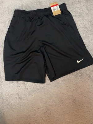 Svarta träningsshorts från Nike - Svarta träningsshorts från Nike med elastisk midja och klassisk vit logga på benet. Lätta och bekväma, perfekta för sport eller gym. Tillverkade i ett syntetmaterial som andas och ger bra rörelsefrihet.