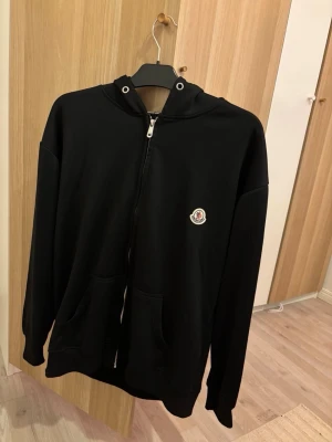 Svart hoodie med dragkedja från Moncler - Svart hoodie från Moncler med dragkedja framtill, två fickor och klassisk huva. På bröstet sitter Monclers ikoniska logotyp i vitt, rött och blått. Hoodien är i mjukt material och har en avslappnad passform, perfekt för en stilren och bekväm look.