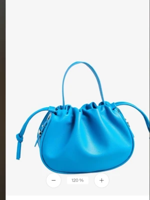 Blå bucket handväska med knytdetaljer - Cool blå handväska i bucket-modell med dragsko och knytdetaljer på sidorna. Väskan har ett kort och långt handtag och är gjord i slätt konstläder. Perfekt för dig som vill ha en färgklick till din outfit.