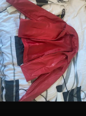 Röd skinnjacka - Snygg röd skinnjacka med klassisk krage och dragkedja framtill. Jackan har en stilren design utan synliga fickor och raka linjer som ger en modern look. Perfekt för dig som vill sticka ut med en färgstark jacka i skinn.