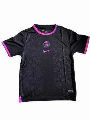 Svart PSG t-shirt från Nike - Snygg svart PSG t-shirt från Nike med lila detaljer på krage och ärmslut. Tröjan har ett diskret blommönster i tyget, PSG-logga och en unik lila Nike swoosh på bröstet. Tillverkad i ett lätt och andningsbart material, perfekt för träning eller streetwear.
