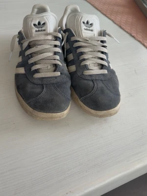 Adidas gråa sneakers i mocka - Snygga Adidas sneakers i grå mocka med vita detaljer och klassiska tre ränder på sidan. Skorna har vit sula och vita skosnören samt Adidas-logga på plös och häl. Perfekta för en avslappnad streetstyle-look.