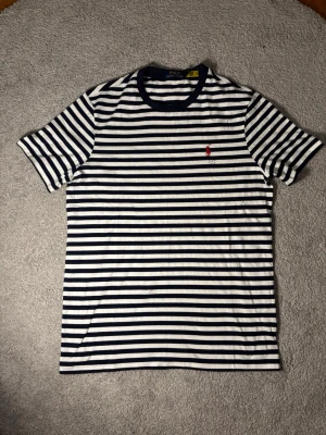 Randig t-shirt från Polo Ralph Lauren - Snygg randig t-shirt från Polo Ralph Lauren i marinblått och vitt. Klassisk passform med korta ärmar och rund hals. Ikonisk röd logga broderad på bröstet. Tillverkad i mjuk bomull som känns skön mot huden.