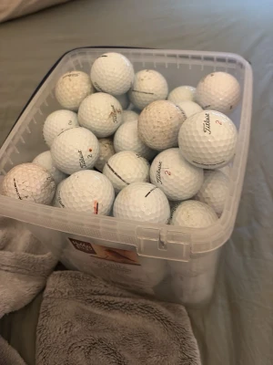 Stor låda med golfbollar - En genomskinlig plastlåda fylld med vita golfbollar, flera av märket Titleist. Perfekt för dig som vill ha många bollar till träning eller spel. Bollarna har klassisk dimple-design och några har siffror och loggor tryckta på sig.