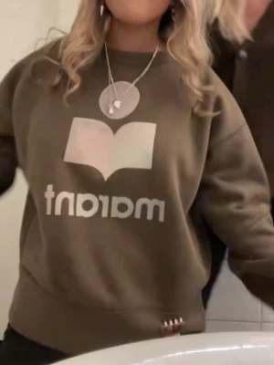 Isabel marant tröja  - Cool olivgrön sweatshirt med ett stort vitt tryck framtill som föreställer en öppen bok och texten 'martnt'. Tröjan har rund halsringning, långa ärmar och ribbade muddar vid ärmslut och nederkant. Perfekt för en avslappnad och trendig look.