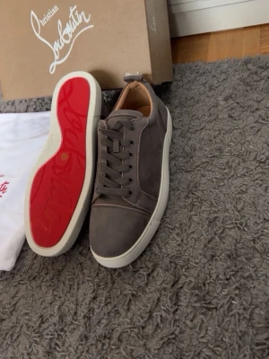 Gråa sneakers från Christian Louboutin - Snygga gråa sneakers från Christian Louboutin med klassisk röd sula och vita kanter. Skorna har en stilren design i mocka och lågt skaft, perfekta för dig som vill ha något exklusivt och trendigt. Matchar enkelt till olika outfits och sticker ut med den ikoniska röda sulan.