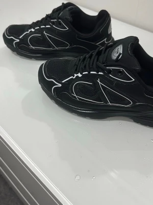 Svarta sneakers med vita detaljer äkta  - Säljer ett par svarta sneakers med vita linjer och CD-logga på hälen. Skorna har en sportig design med mesh och syntetmaterial, samt svarta snören och en tjock sula för extra komfort. Perfekta för dig som gillar en clean och modern look.