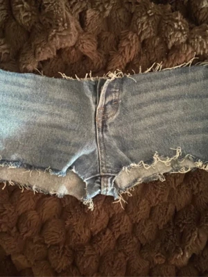 Low rise jeansshorts med fransig kant - Säljer ett par blå jeansshorts med rå, fransig kant och låg midja. Shortsen har klassisk femficksdesign och är perfekta för varma sommardagar. De är gjorda i bomull och har en avslappnad, lite tight passform. Dom är väldigt low rise så var beredd 