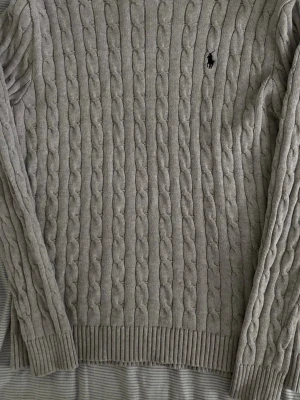 Grå kabelstickad tröja Polo Ralph Lauren - Kabelstickad tröja i ljusgrått från Polo Ralph Lauren med klassisk svart broderad logga på bröstet. Tröjan har långa ärmar, ribbade muddar och rund hals. Perfekt för lager på lager och en stilren look.