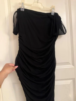 Svart offshoulder meshklänning - Snygg svart kortklänning med offshoulder-design och meshdetaljer på ärmarna. Klänningen har draperad effekt längs hela kroppen och är figurnära. Perfekt för dig som vill ha en trendig och stilren look.
