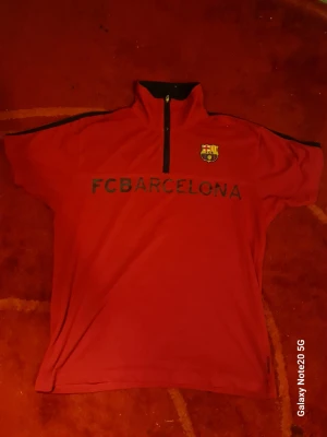 Röd FCB Barcelona topp med dragkedja - Röd topp med svart dragkedja och hög krage från FCB Barcelona. Tryckt klubbmärke på bröstet och texten 'FC BARCELONA' över magen. Svarta detaljer längs axlarna. Perfekt för dig som älskar fotboll och vill visa ditt stöd för laget.