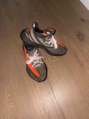 Yeezy Boost 350 V2 - Säljer ett par Yeezy Boost 350 V2 med orange stripe och texten SPLY-350 på sidan. Skorna har en stickad ovandel i grått och svart med vita skosnören och svart sula. Perfekta för dig som gillar streetwear och vill sticka ut.
