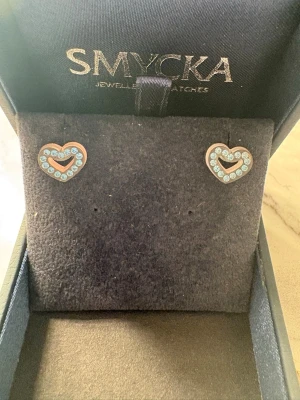 Tre par örhängen från Smycka - Säljer tre par örhängen från Smycka. Ett par är hjärtformade i guldton med ljusblå stenar, och det andra paret är små fjärilar i guldton med glittrande stenar. Perfekta för att addera lite extra bling till din stil.
