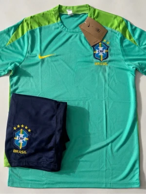 Brasilien träningsset Nike grön/blå - Snyggt träningsset från Nike med Brasilien CBF-logga. Tröjan är ljusgrön med limegröna axelpartier och gula detaljer, samt rund halsringning. Tillhörande shorts i mörkblått med matchande logga. Perfekt för fotbollsträning eller sport.